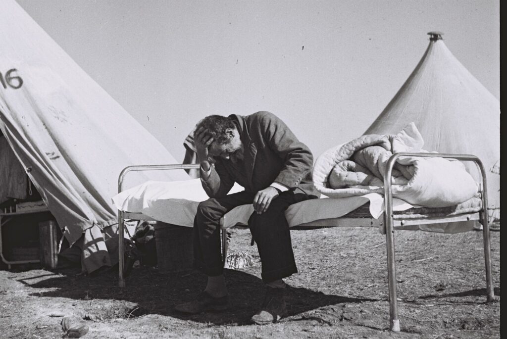 עולה חדש יושב על מיטתו מחוץ לאוהל במחנה עולים בתל אביב - פין הנס (חיים)